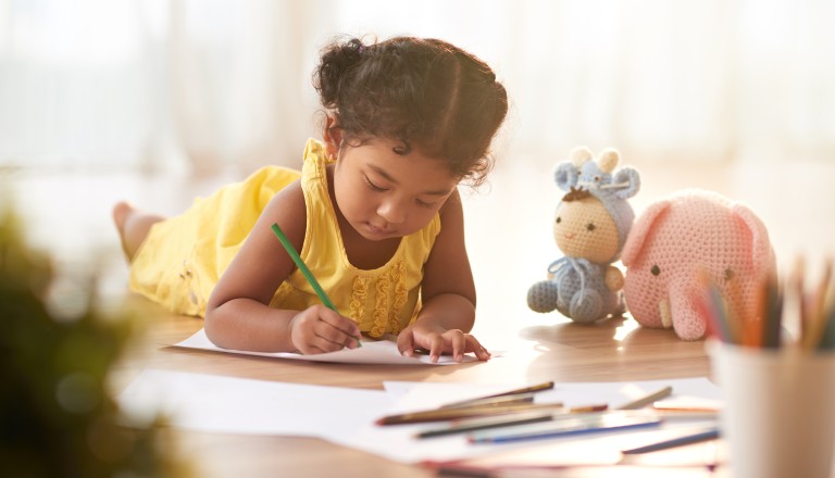 Concentrated toddler in yellow dress coloring picture with pencils while lying on floor, blurred background