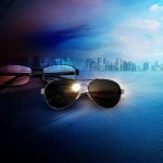 Stylized photo of a pair of glasses and a pair of sunglasses in the foreground, with a city skyline and sky in the background. The left side of the photograph is dark and the right side of the photograph is bright.